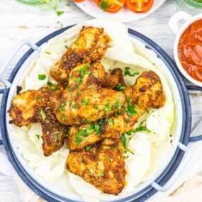 Golden-brown chicken wings garnished with herbs, served on cabbage leaves in a round dish, with a side of red sauce and cherry tomatoes nearby.