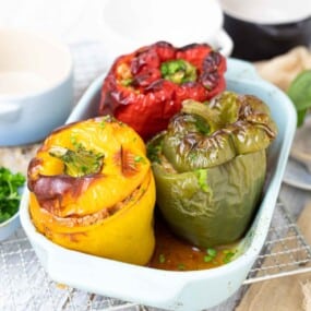 Three roasted stuffed bell peppers in red, yellow, and green, placed in a light blue baking dish on a wire rack.