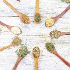 Wooden spoons arranged in a circle on a white wooden surface, each containing different dried herbs and spices.
