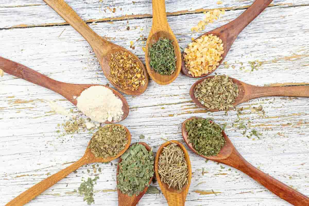 Wooden spoons arranged in a circle, each holding different dried herbs and spices on a weathered white wooden surface.
