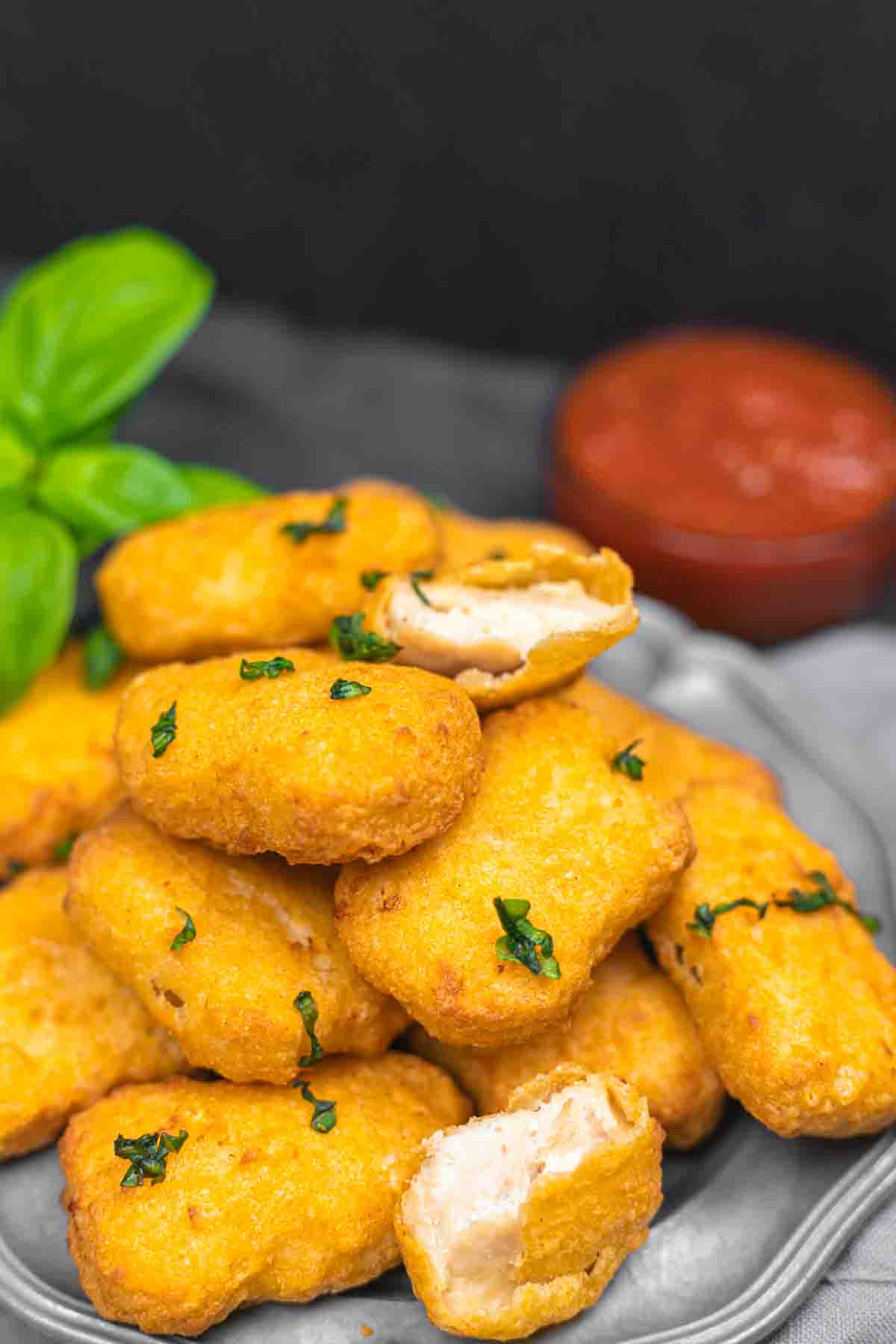 A pile of golden-brown, rectangular nuggets garnished with herbs on a plate, with a bowl of red sauce and basil leaves in the background.