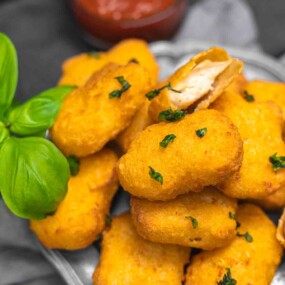 A pile of golden, crispy chicken nuggets garnished with herbs, with one nugget bitten into, on a plate next to basil leaves and a bowl of dipping sauce in the background.