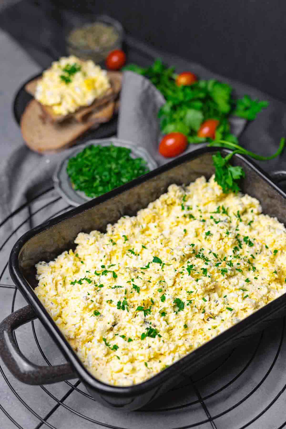 A baking dish filled with scrambled eggs topped with herbs. In the background, there are slices of bread with eggs, herbs, tomatoes, and a small bowl of chopped greens.
