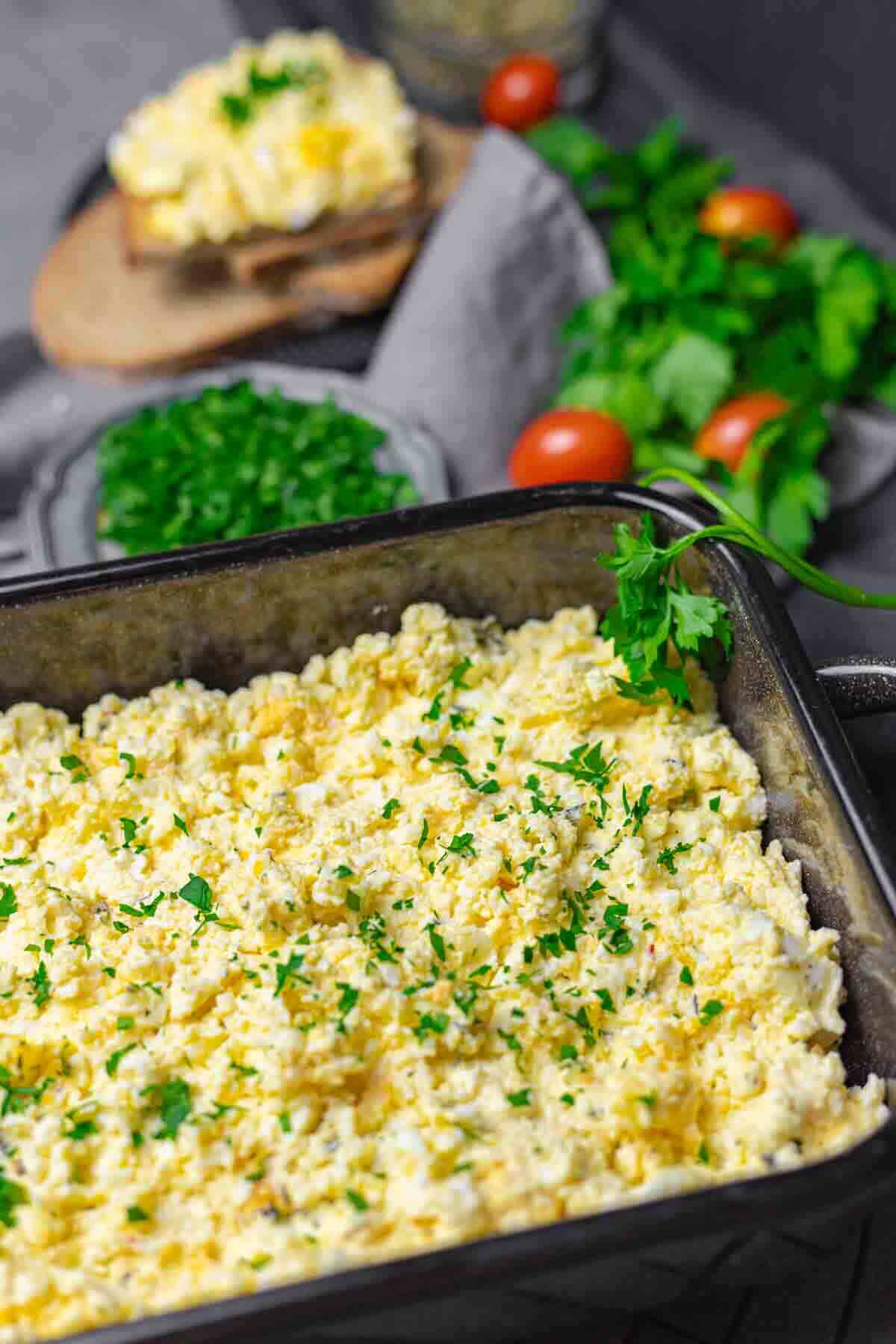 A baking dish filled with scrambled eggs topped with chopped herbs. Fresh herbs and cherry tomatoes in the background.