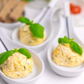 Three white spoons with scoops of cheese topped with basil leaves, placed on a white surface.