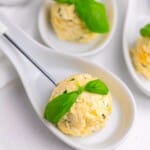 A white spoon holds a scoop of herbed yellow butter, garnished with a green basil leaf, with similar dishes blurred in the background.