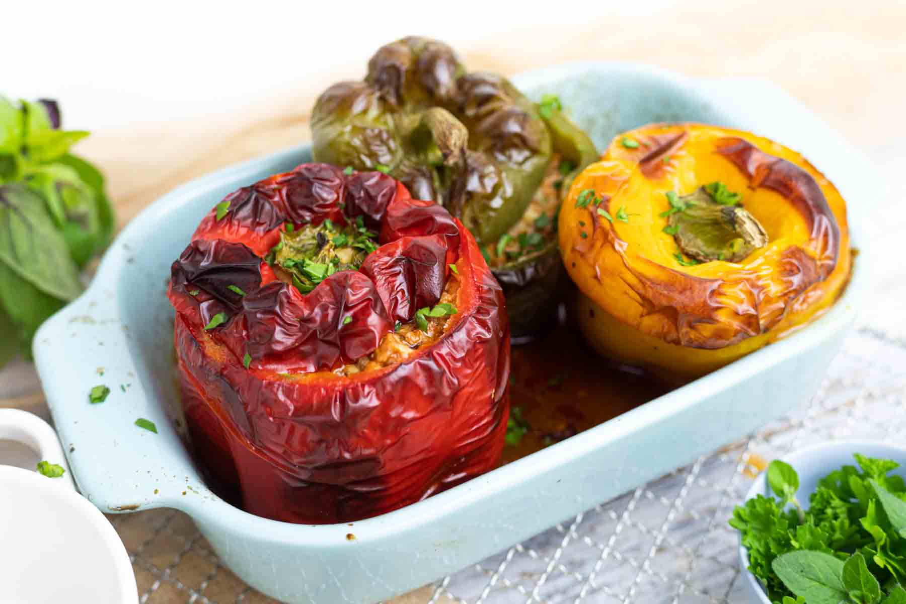 Three stuffed bell peppers, one red, one green, and one yellow, cooked and garnished with herbs, served in a light blue baking dish.