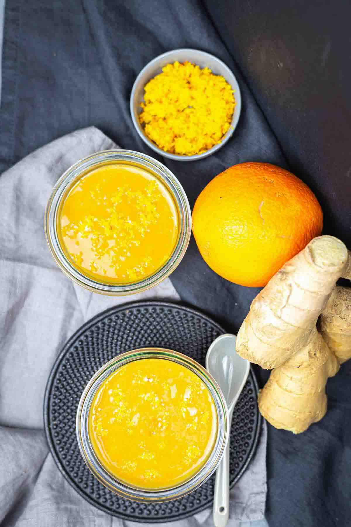 Two jars of orange-colored smoothie with grated zest in a small bowl, an orange, and ginger root on a dark surface with a gray cloth and a spoon.