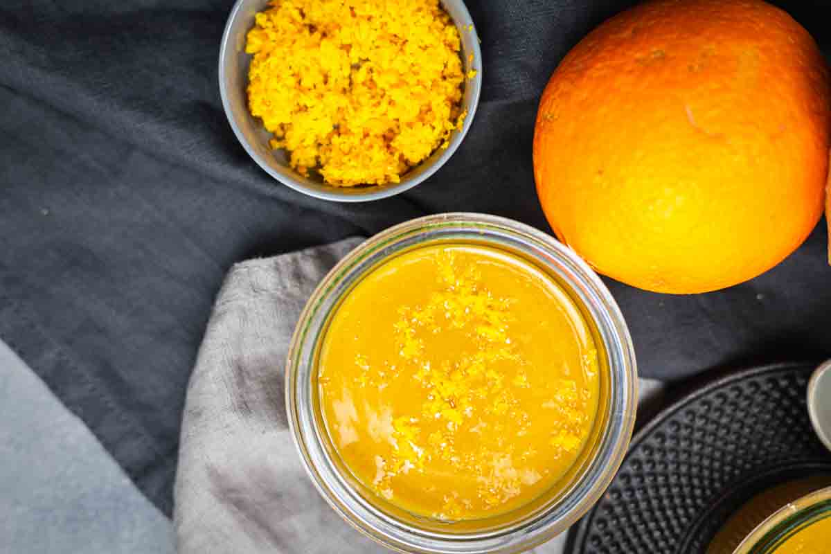 Jar of orange sauce next to a small bowl of grated orange zest and a whole orange on a gray cloth background.