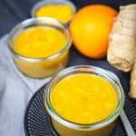 Two glass jars of orange-colored turmeric paste are on a table, accompanied by fresh ginger, an orange, and a small bowl with turmeric powder.