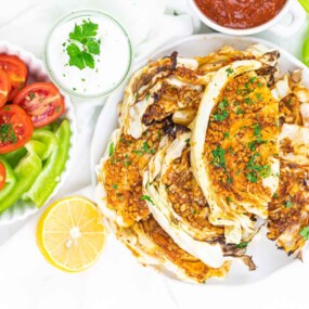 Plate of cabbage rolls filled with a brown stuffing, garnished with cilantro. Accompanied by sliced tomatoes, green peppers, dip, salsa, and a lemon half on a white background.