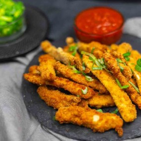 A plate of breaded and fried chicken fries garnished with herbs, served with a bowl of red dipping sauce and a green dip in the background.