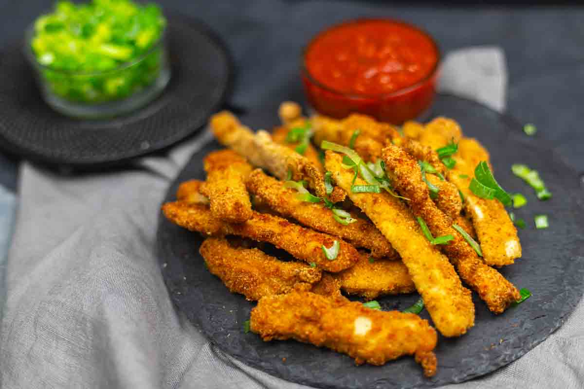 A plate of breaded and fried chicken fries garnished with herbs, served with a bowl of red dipping sauce and a green dip in the background.