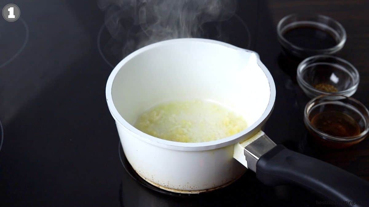 A white saucepan on a stovetop with garlic sizzling in oil. Three small glass bowls containing spices are placed on the right side. Steam is rising from the pan.