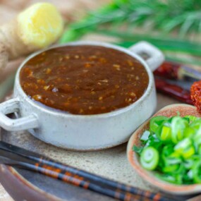 A bowl of brown sauce next to a dish of sliced green onions, red chili peppers, and a ginger root on a plate.