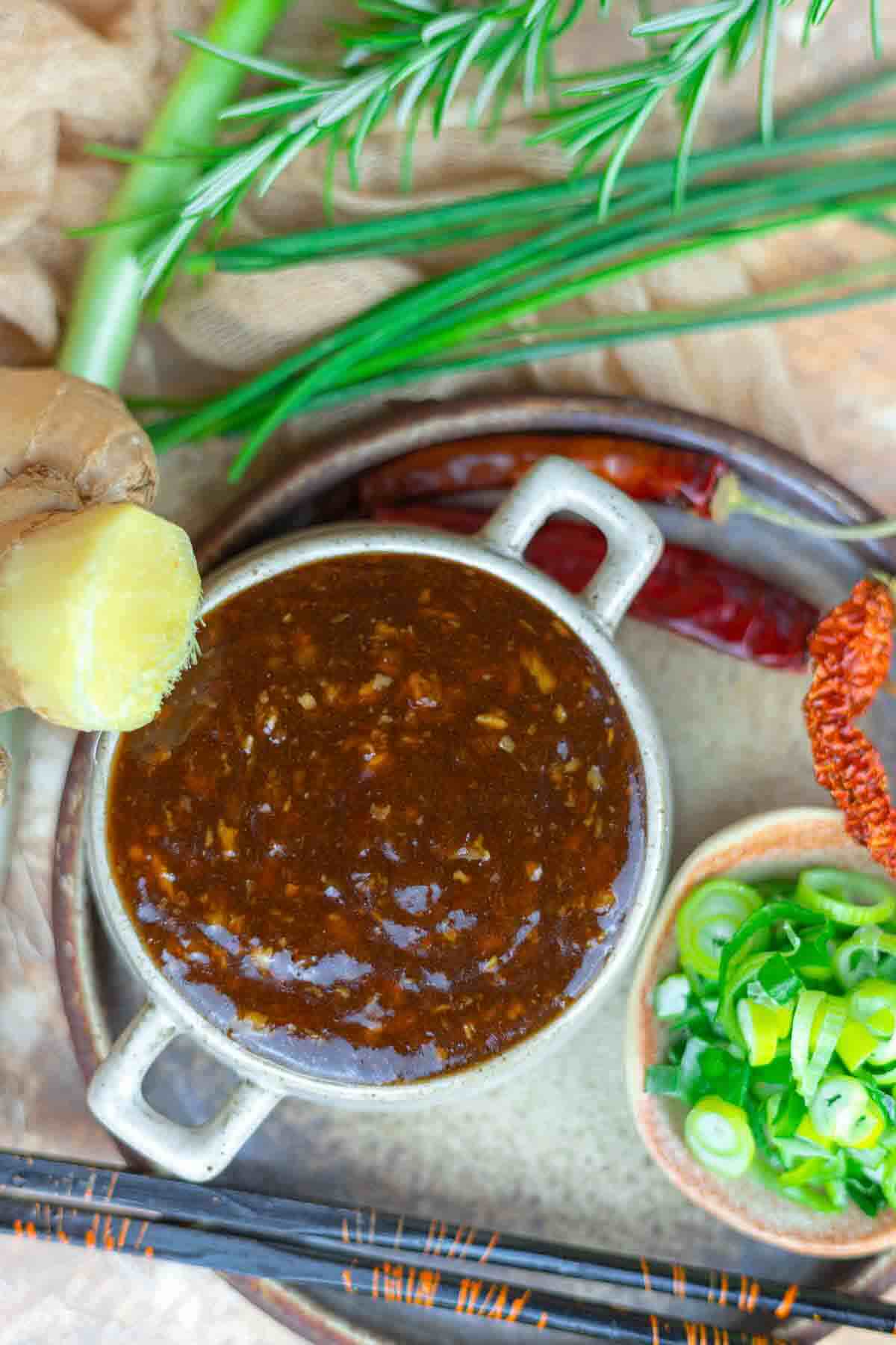 Bowl of dark sauce surrounded by fresh ginger, scallions, rosemary, chives, dried peppers, and chopsticks on a textured surface.