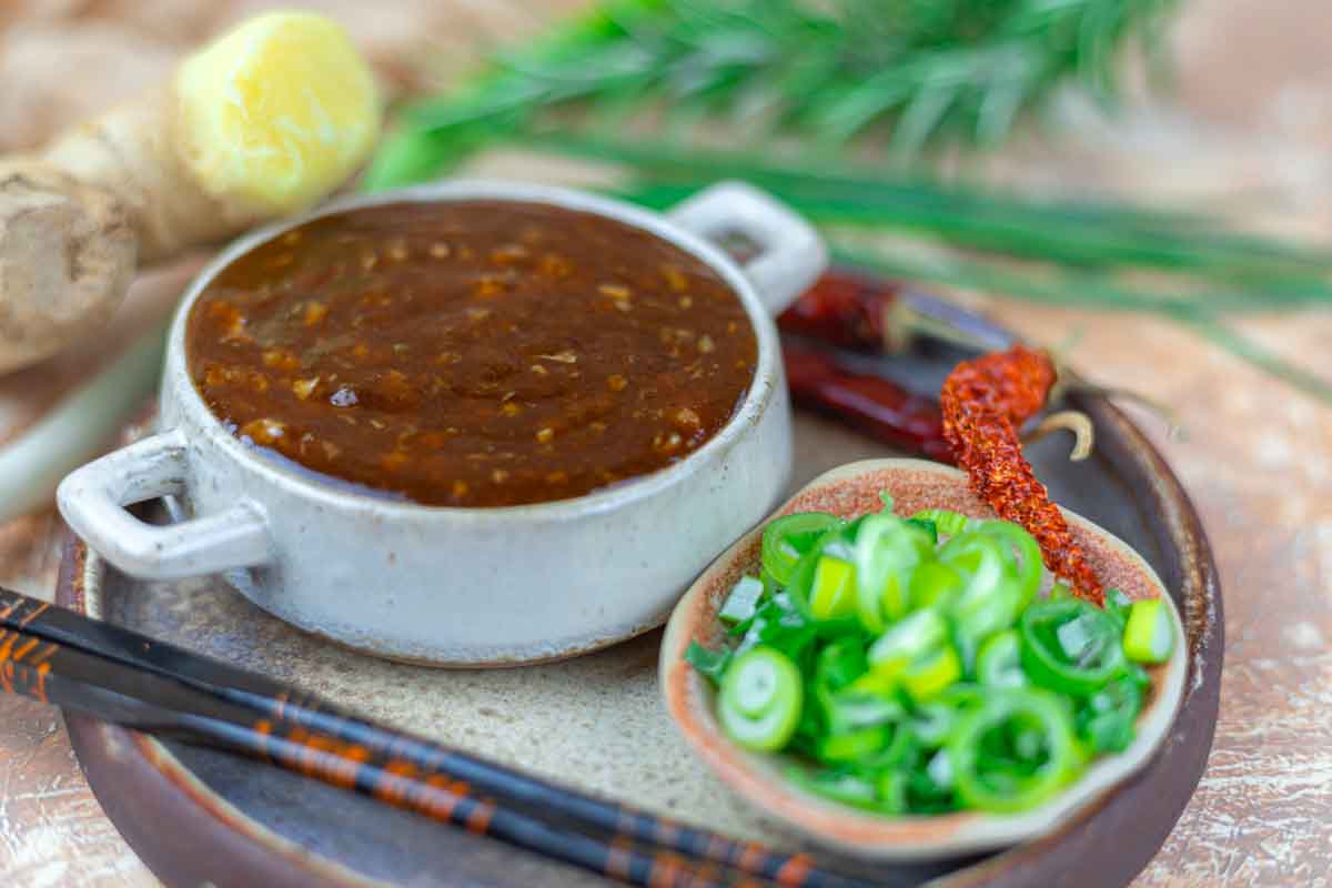 Bowl of Asian BBQ sauce next to a dish of sliced green onions and a dried chili pepper, with chopsticks, ginger, and herbs in the background.