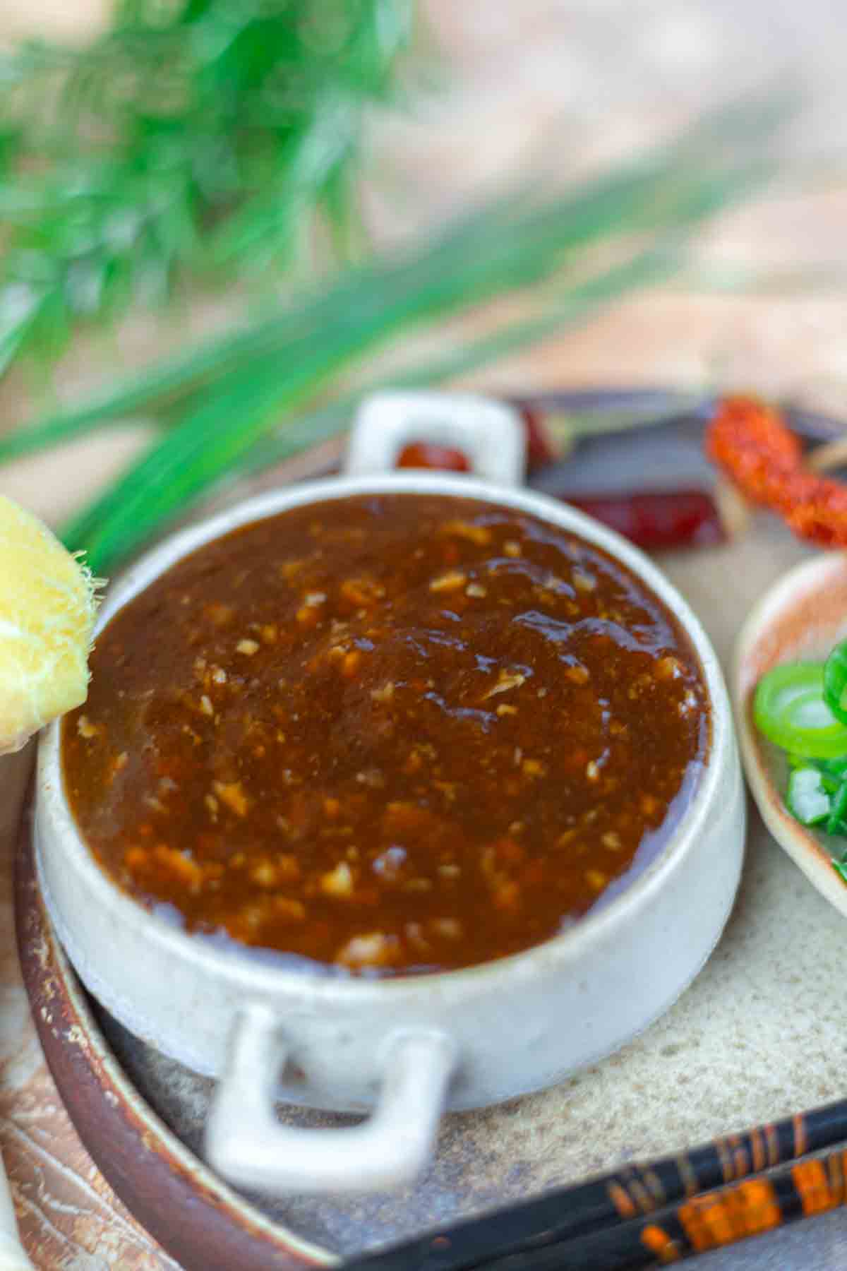 A ceramic dish filled with thick, brown sauce, surrounded by green onions, red chili peppers, and a piece of ginger on the side.