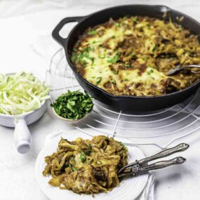 A skillet with melted cheese pasta beside small bowls of shredded cabbage and herbs. A serving is plated on a white dish with a fork and spoon.