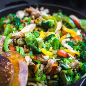 A skillet filled with a colorful stir-fry, including broccoli, bell peppers, and chicken, with a wooden spatula on the side.