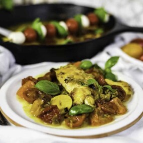 A plate with baked fish fillet topped with sauce, surrounded by cherry tomatoes and basil. A pan with skewered tomatoes and cheese is in the background.