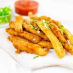 A pile of breaded and fried chicken strips garnished with chopped greens on a white surface. Small bowls of chopped green onions and red sauce in the background.