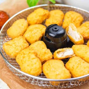 A wire basket filled with golden-brown chicken nuggets, some bitten, with a small cup of red dipping sauce beside it.