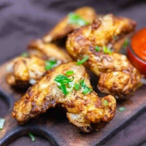 Crispy fried chicken wings garnished with chopped green onions, served on a wooden tray with a side of red dipping sauce.