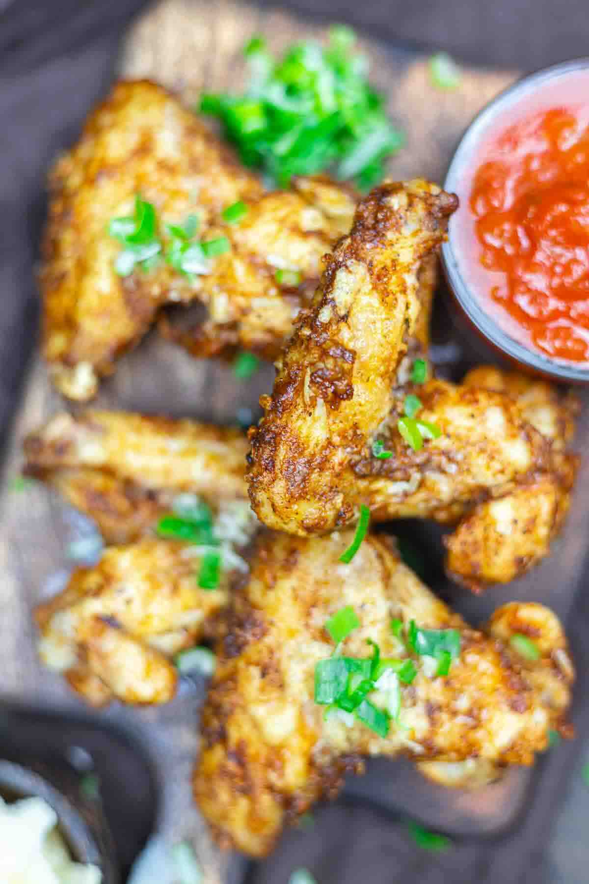 Three seasoned fried chicken wings with garnish are on a wooden board, accompanied by a red dipping sauce and chopped green herbs.