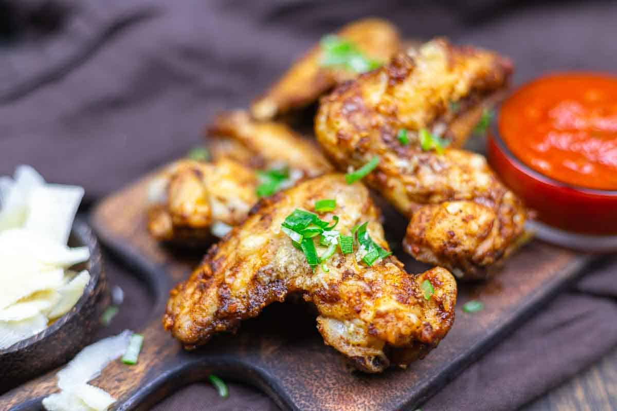 Fried chicken wings garnished with chopped green onions on a wooden board, accompanied by a bowl of red sauce and a side of sliced cheese.