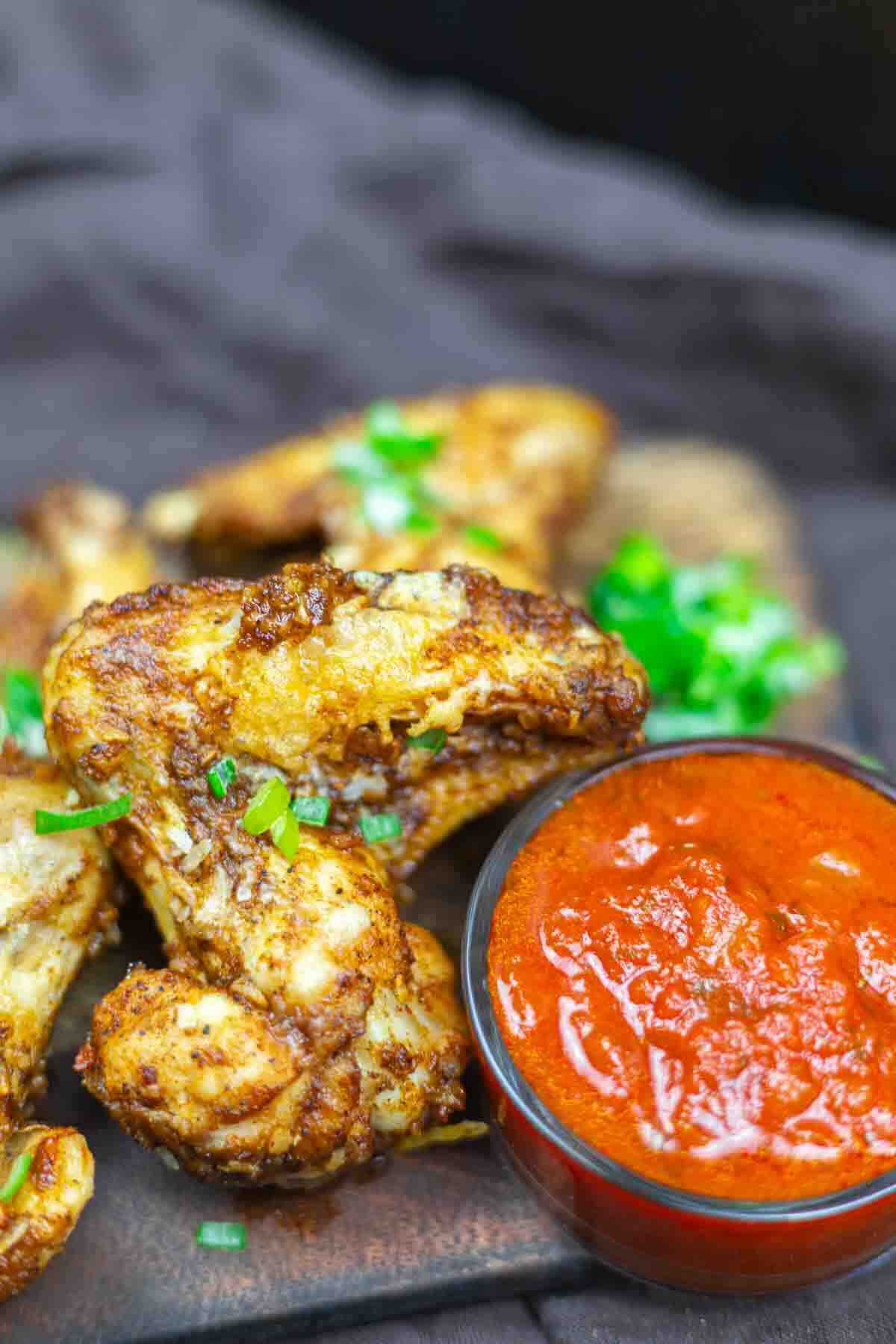 Crispy chicken wings with a garnish of chopped green herbs next to a small bowl of red sauce.