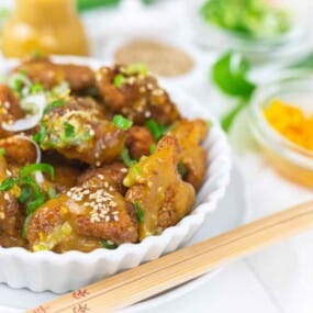 A white dish filled with orange chicken topped with sesame seeds and green onions, surrounded by small bowls of toppings and a pair of chopsticks.