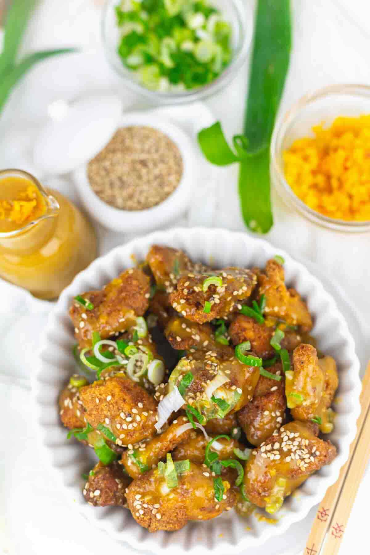 A bowl of crispy fried chicken pieces garnished with sesame seeds and green onions, surrounded by ingredients like chopped scallions, sesame seeds, and sauce in small bowls.