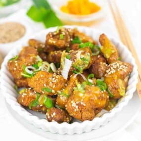 A white bowl filled with crispy fried chicken pieces topped with sesame seeds, green onions, and drizzled with a light sauce. Chopsticks and small dishes of spices are in the background.