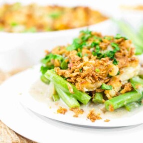 A plate of green bean casserole topped with crispy onions and chopped herbs, with more casserole in a dish in the background.