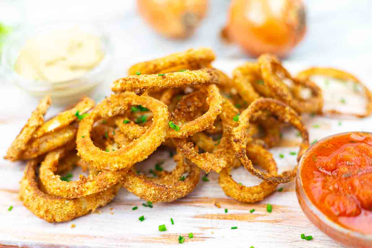 A pile of crispy onion rings garnished with chives, accompanied by small dishes of dipping sauces, are shown on a light wooden surface.