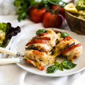 Grilled stuffed chicken breast with tomatoes and cheese, garnished with basil leaves, served on a white plate with salad and fresh vegetables in the background.