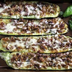 Four stuffed zucchini halves topped with melted cheese and herbs are arranged in a baking dish, garnished with fresh basil leaves.
