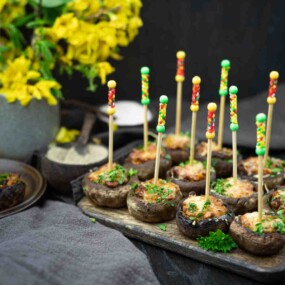 Stuffed mushrooms with decorative skewers on a wooden platter, garnished with parsley. Yellow flowers in a vase are in the background.