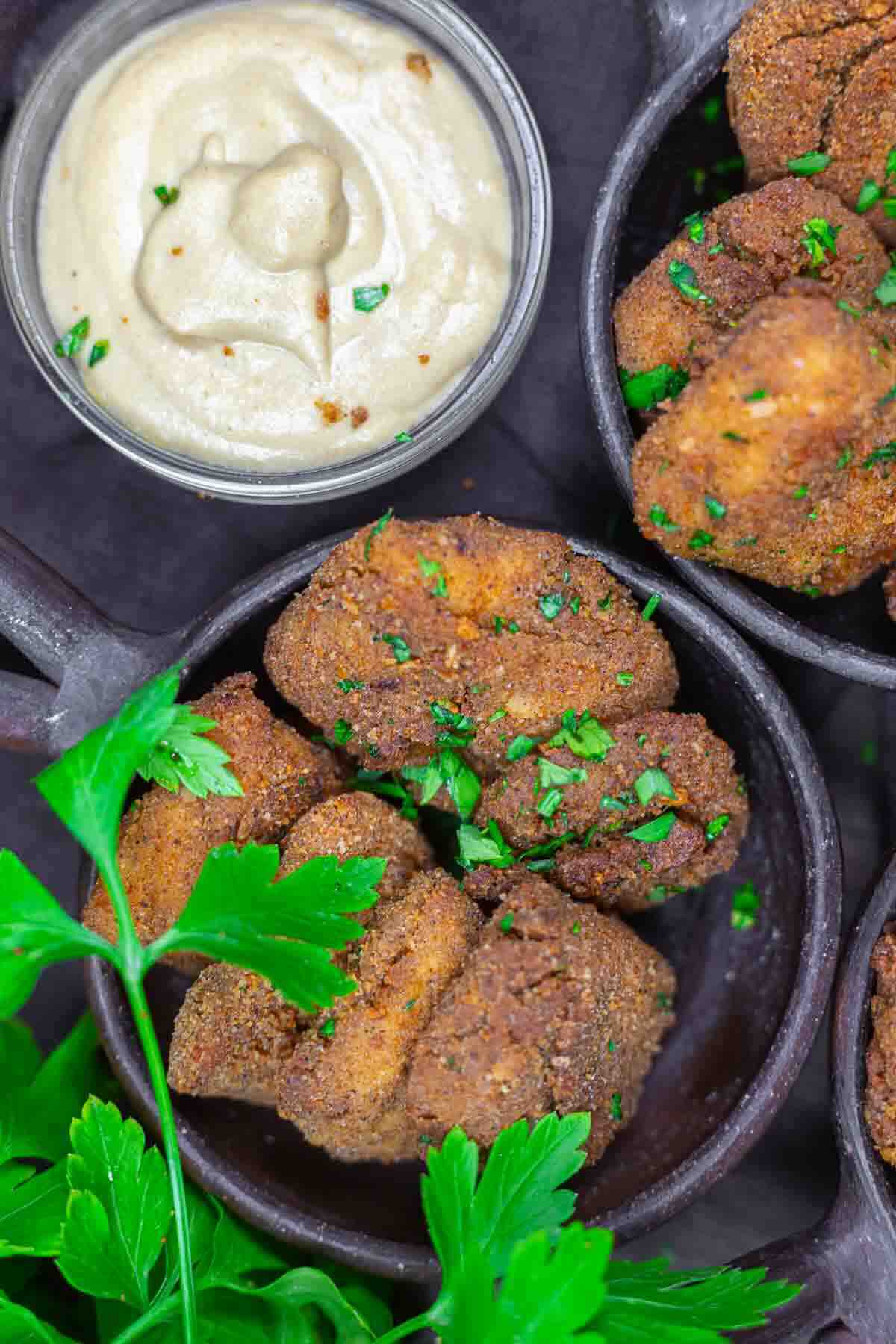 Close-up of crispy, breaded vegan nuggets in a black bowl, garnished with chopped parsley, next to a small bowl of creamy dipping sauce. Fresh parsley leaves are also visible.