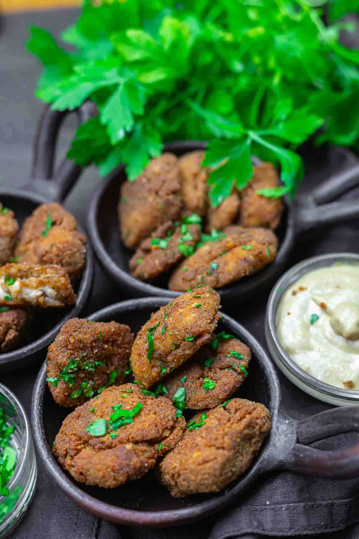 Three black bowls filled with falafel patties, garnished with chopped herbs, are arranged next to a bowl of dipping sauce and a bunch of fresh parsley.