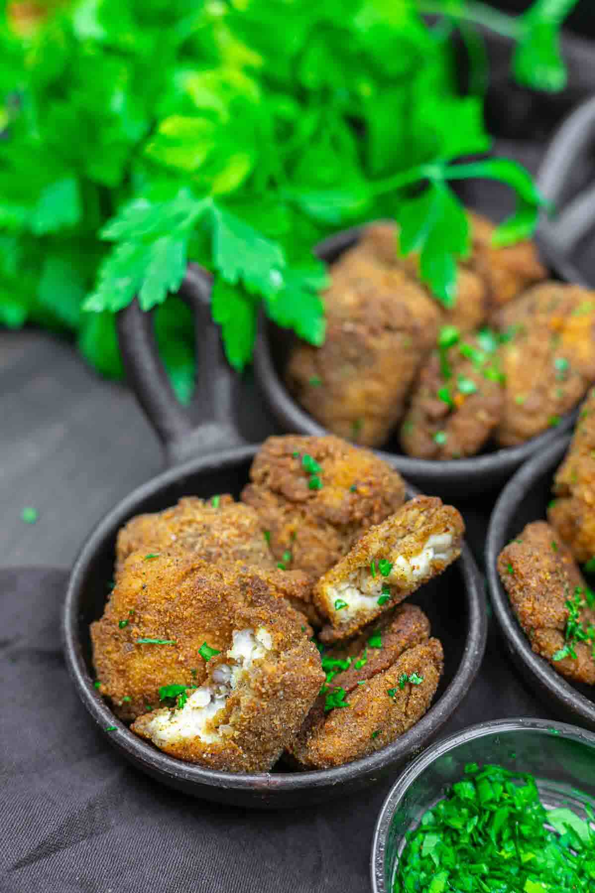 Breaded and fried pieces of paneer arranged in small black bowls, garnished with chopped parsley, with fresh parsley in the background.