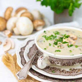 A bowl of creamy mushroom soup garnished with herbs, placed on a patterned saucer with a spoon, and a plate of fresh mushrooms in the background.