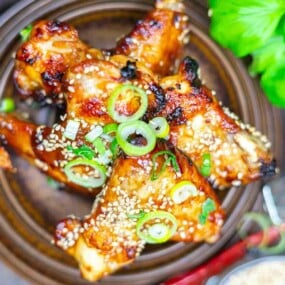 Grilled chicken drumsticks topped with sesame seeds and sliced green onions, served in a brown bowl with leafy greens in the background.