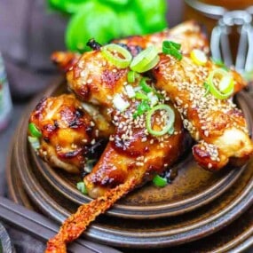 Oven baked BBQ wings garnished with sesame seeds and sliced green onions, served on a brown ceramic plate with decorative items in the background.