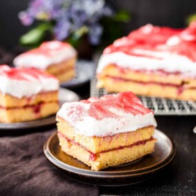 Three slices of layered cake with white frosting and red topping are displayed on plates and a cooling rack with a dark background.