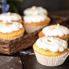 Cupcakes with white frosting sit on a dark wooden table. A woven basket holds more cupcakes in the background, next to a dark cloth.