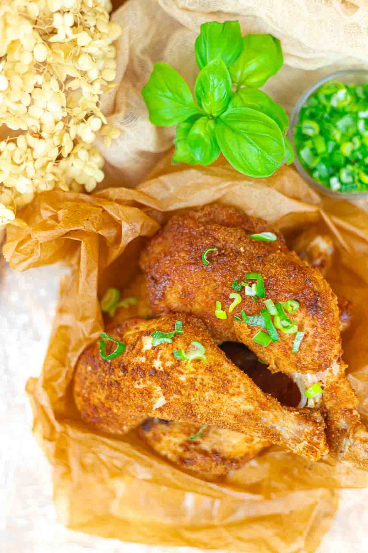 Two seasoned baked chicken drumsticks garnished with chopped green onions, served on brown parchment paper with fresh basil and sliced green chili on the side.