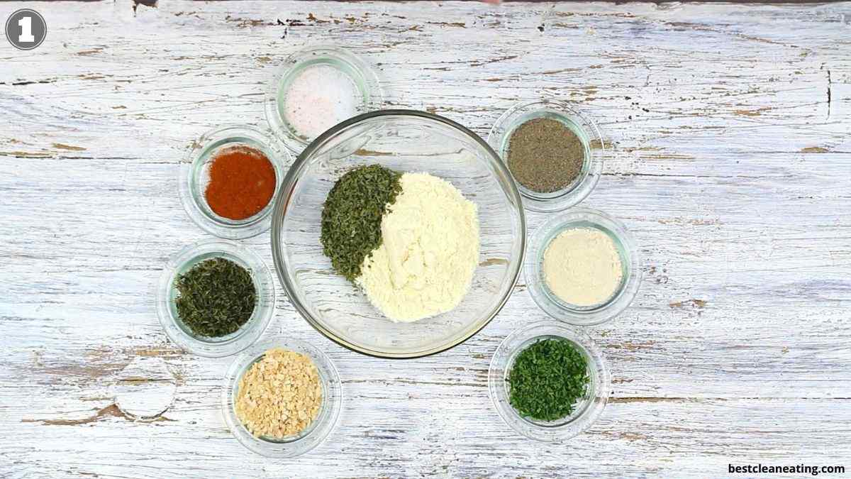 A glass bowl with powder and dried herbs is surrounded by eight small bowls with various spices and seasonings on a white wooden surface.