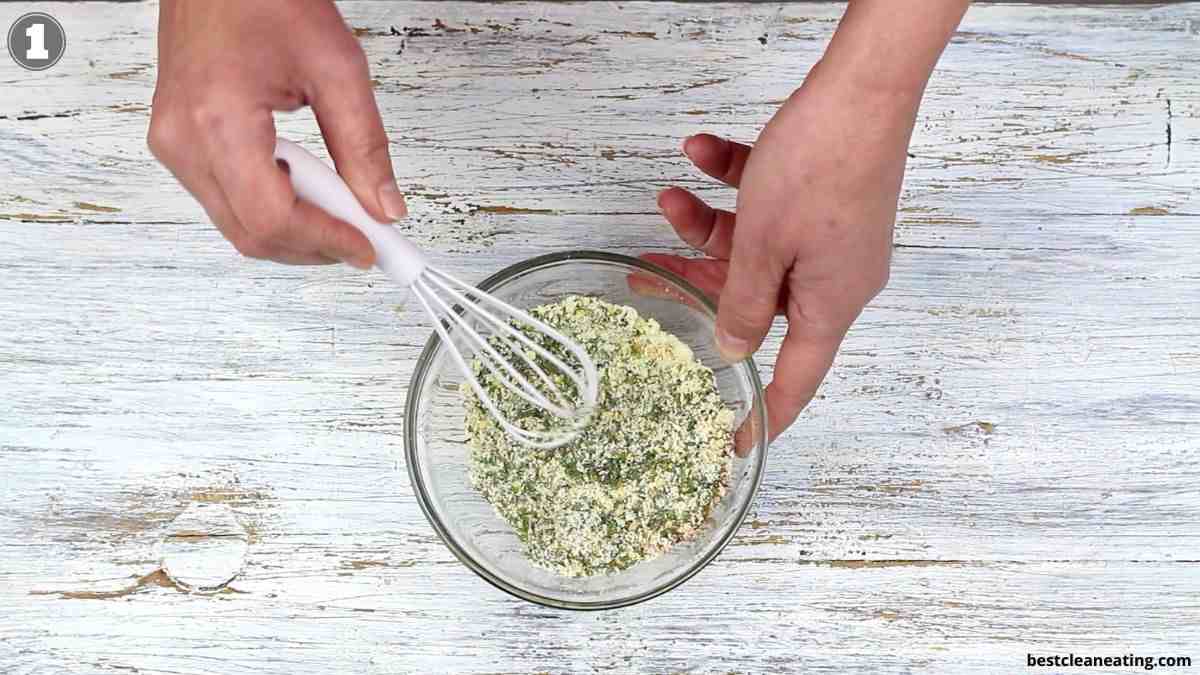 A person uses a white whisk to mix dry ingredients in a glass bowl on a white wooden surface.